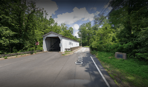 A covered bridge surrounded by lush greenery, with a road leading to it and a stop sign visible.