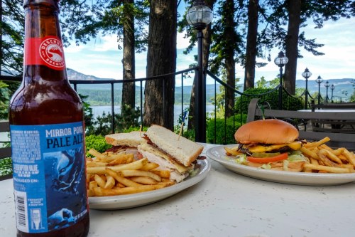 A scenic outdoor table with a sandwich, burger, fries, and a bottle of pale ale, surrounded by trees and a lake view.