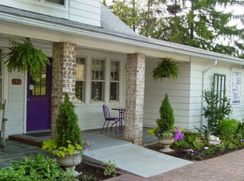 Charming house entrance with purple door, potted plants, and hanging ferns, surrounded by a brick walkway.