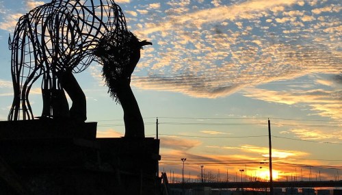 Silhouette of a wire sculpture against a colorful sunset with clouds and power lines in the background.