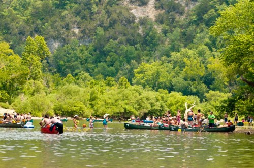 A busy river scene with people kayaking and swimming, surrounded by lush green trees and hills.