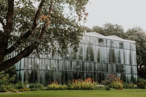 A modern glass greenhouse surrounded by lush greenery and colorful flowers under a cloudy sky.
