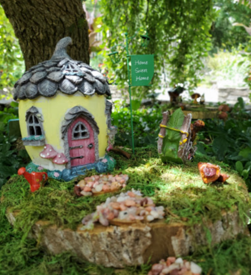A whimsical fairy house with a thatched roof, surrounded by moss, mushrooms, and a "Home Sweet Home" sign.