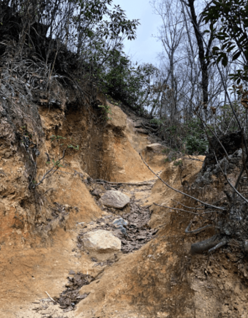 A narrow, eroded trail winding through a hillside with exposed soil and sparse vegetation.