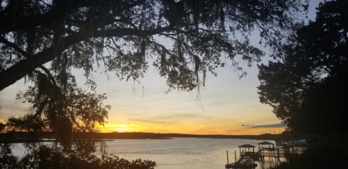 A serene sunset over a calm lake, framed by trees and Spanish moss, with a dock visible in the distance.