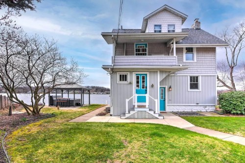 A charming gray house with blue accents, overlooking a lake, surrounded by green grass and trees.