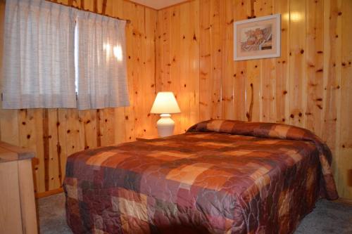 Cozy bedroom with wooden walls, a patterned quilted bedspread, a lamp, and a window with sheer curtains.