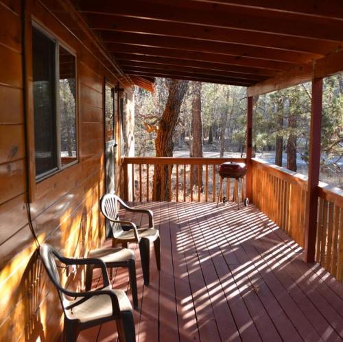 A wooden porch with two chairs and a grill, surrounded by trees and sunlight filtering through.
