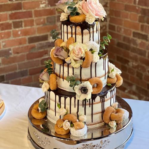 A three-tiered cake adorned with flowers and donuts, featuring chocolate drizzle, set against a brick wall backdrop.