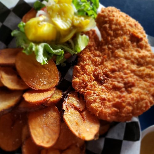 A plate featuring a crispy fried chicken breast, seasoned potato wedges, and a side of lettuce and pickles.