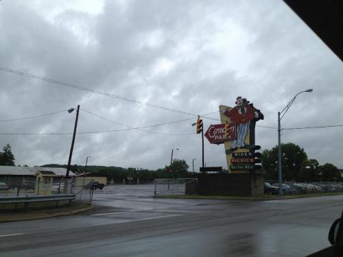A vintage amusement park sign on a cloudy day, with empty parking and a few buildings in the background.