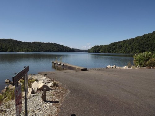 A calm lake surrounded by green hills, with a boat ramp and clear blue sky above.