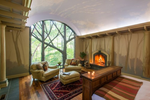 Cozy bedroom with a fireplace, plush seating, and a large window featuring a serene tree mural. Warm, inviting atmosphere.