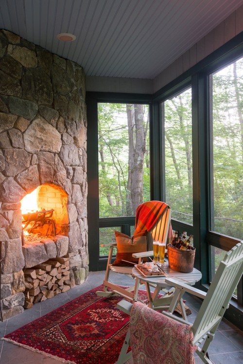 Cozy screened porch with a stone fireplace, chairs, a rug, and a view of trees outside. Warm and inviting atmosphere.