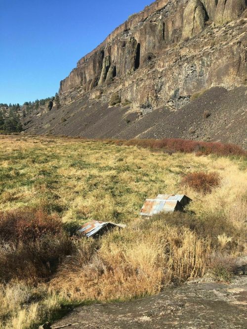 A weathered metal structure lies in a grassy field, with rocky cliffs rising in the background under a clear blue sky.