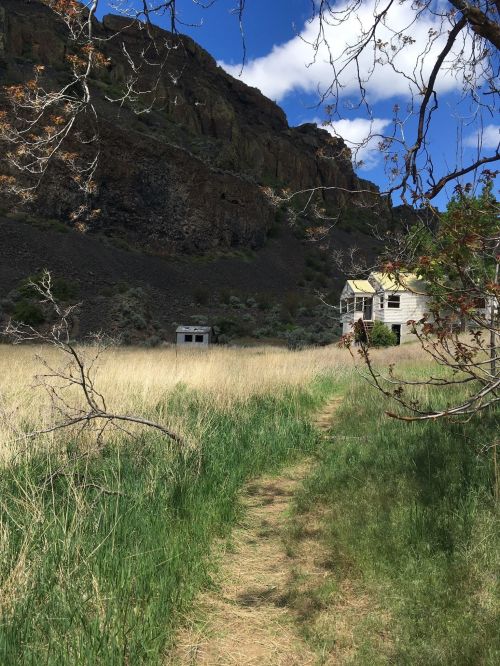A grassy path leads to two abandoned buildings, surrounded by rocky cliffs and a blue sky with scattered clouds.