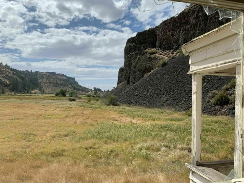 A view of a grassy field bordered by rocky cliffs under a partly cloudy sky, seen from a rustic wooden structure.
