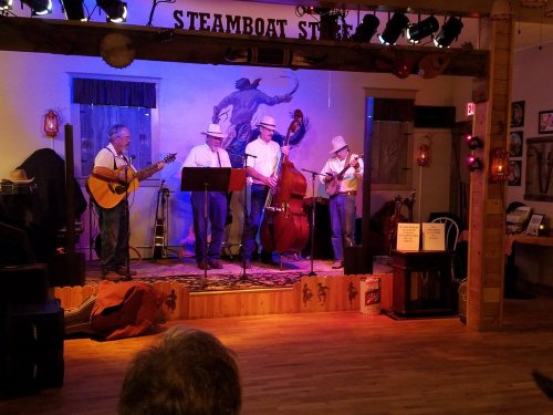 A band performs on stage with a guitar, bass, and fiddle in a cozy venue decorated with western-themed art.