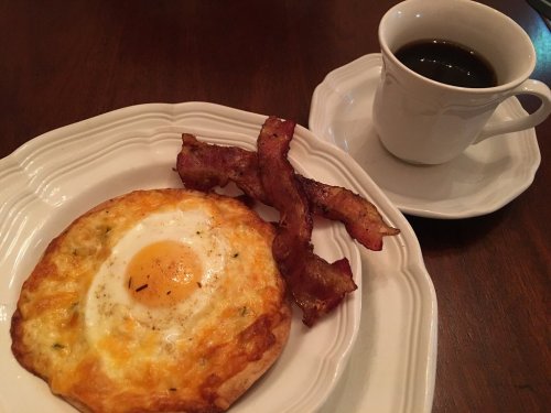 A plate with a cheesy egg dish and crispy bacon, alongside a cup of black coffee on a wooden table.