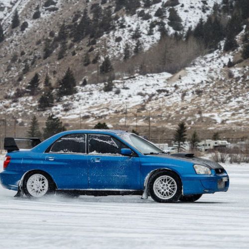 A blue car drifts on a snowy landscape with mountains in the background. Snow is swirling around the tires.