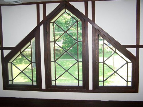 Triangular wooden windows with diamond-patterned panes, set in a white wall, overlooking a green outdoor area.