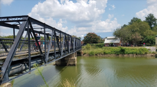 A metal bridge spans a green river, with trees and a building visible in the background under a partly cloudy sky.