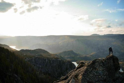 A person stands on a rocky outcrop, overlooking a vast landscape of mountains and a lake under a cloudy sky.