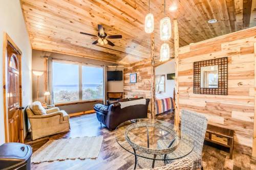 Cozy living room with wooden walls, a ceiling fan, and a view of the outdoors through large windows.
