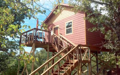 A red and yellow treehouse with a deck, surrounded by trees and blue sky. Stairs lead up to the entrance.