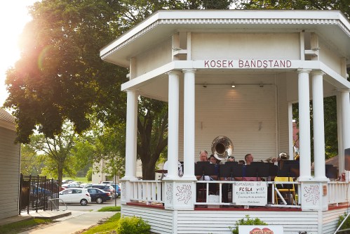 A band performs in a gazebo labeled "Kosek Bandstand" with trees and parked cars in the background.