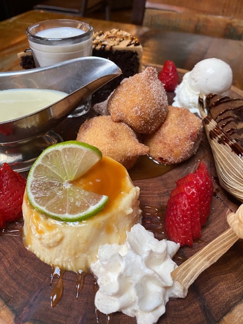 A wooden platter with various desserts: flan, chocolate cake, fried dough balls, strawberries, and ice cream.