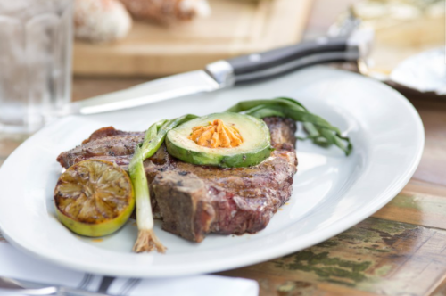 A grilled steak topped with avocado and a slice of lime, garnished with green onions on a white plate.