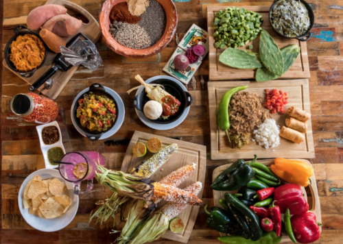 A vibrant spread of fresh vegetables, herbs, and dishes, including sweet potatoes, corn, and various toppings on wooden boards.