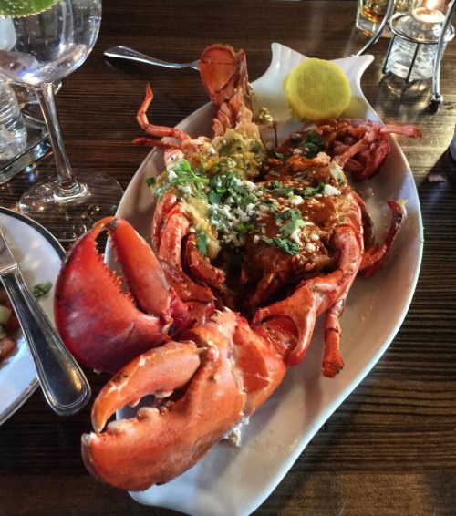 A whole lobster served on a white platter, garnished with herbs, lemon, and a side of sauce.