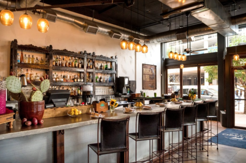 A stylish restaurant bar with a wooden shelf of liquor, bar stools, and warm lighting, featuring cacti and sunflowers.