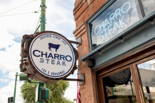 Sign for Charro Steak restaurant, featuring a cow graphic and the text "Charro Steak" against a brick building.