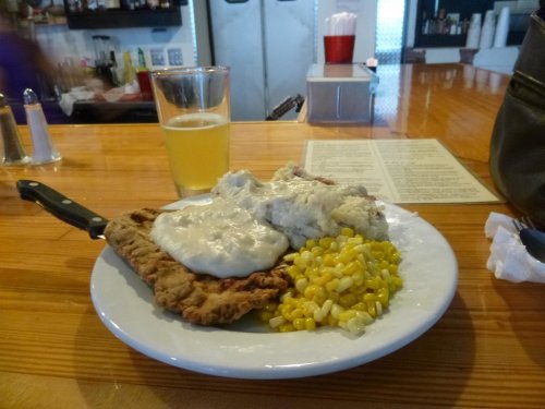 A plate of food featuring fried chicken with gravy, mashed potatoes, corn, and a glass of beer on a wooden table.
