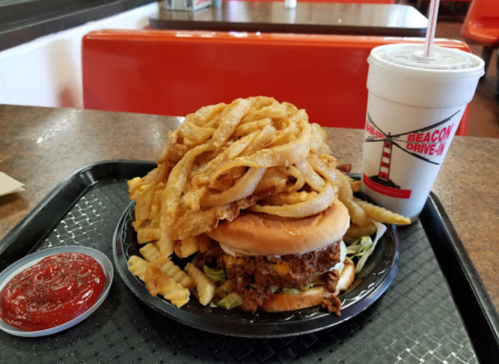 A large burger topped with lettuce and sauce, surrounded by crispy onion rings and fries, with a drink on the side.