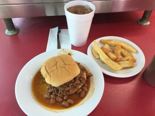 A plate with a sandwich topped with chili, beans, and a side of fries, accompanied by a large drink on a red table.