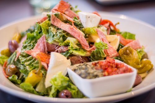 A colorful salad with greens, meats, cheese, olives, and a side of dressing in a white bowl.