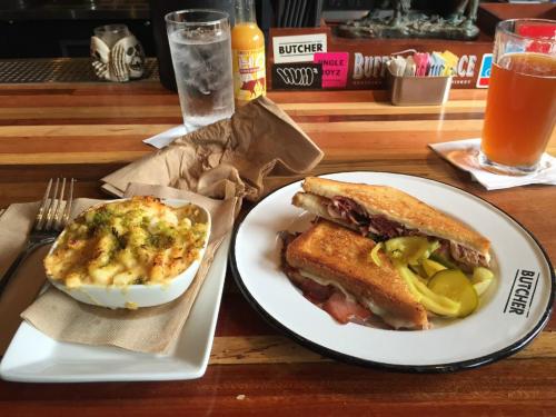 A plate with a grilled sandwich and pickles next to a bowl of mac and cheese, with drinks in a rustic bar setting.