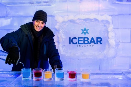 A man in winter attire mixes colorful drinks at ICEBAR Orlando, surrounded by ice decor and a logo in the background.