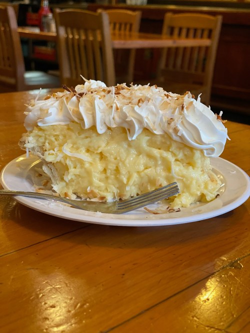 A slice of coconut cream pie topped with whipped cream and toasted coconut, served on a white plate.