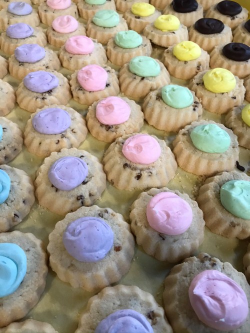 A close-up of colorful, frosted mini cakes arranged in rows, featuring pastel shades of pink, blue, green, and yellow.