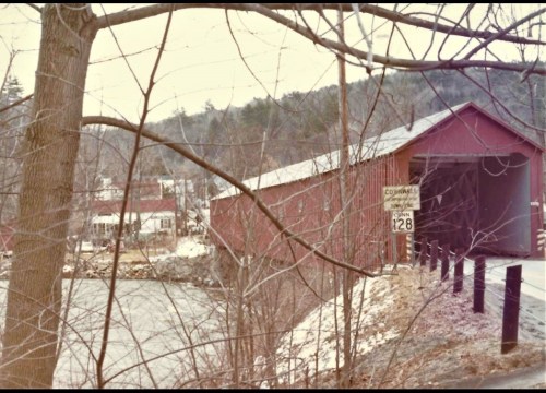 A red covered bridge over a river, surrounded by bare trees and a snowy landscape, with mountains in the background.