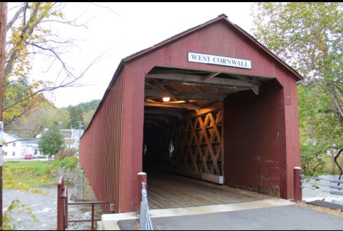 A red covered bridge labeled "West Cornwall" with wooden beams, surrounded by trees and a small town in the background.