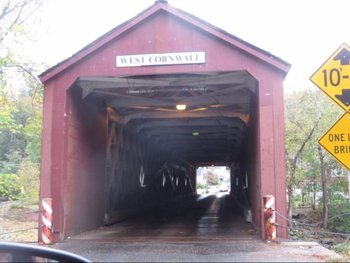 A red covered bridge with a sign reading "West Cornwall," viewed from the entrance, with a warning sign nearby.