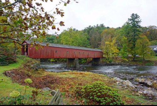 A red covered bridge spans a river, surrounded by autumn foliage and trees in a serene landscape.