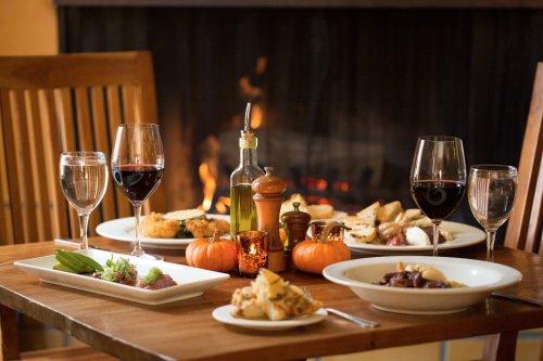 A cozy dinner setting with plates of food, wine glasses, and candles, near a warm fireplace. Small pumpkins are on the table.