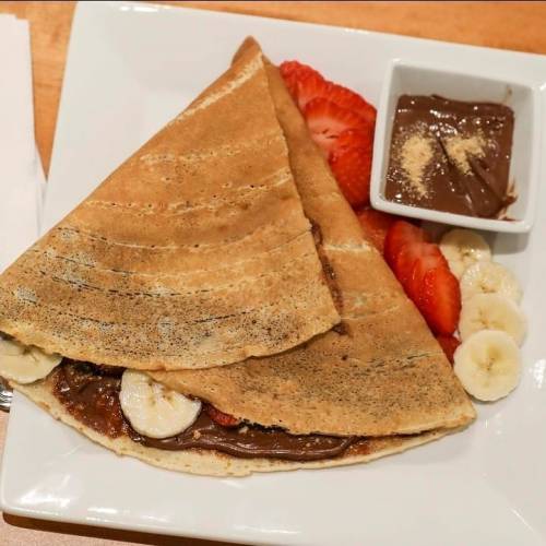 A folded crepe filled with chocolate spread, sliced bananas, and strawberries, served on a white plate with a small bowl of chocolate.
